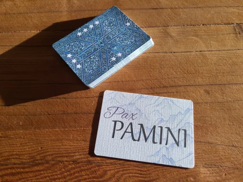 The Pax Pamini title card beside a deck of its blue patterned card backs on a wooden table.