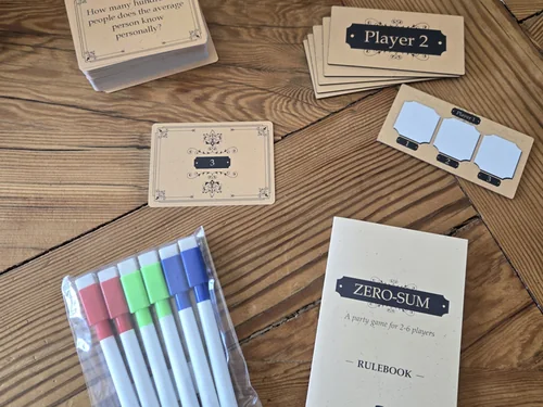 Zero-Sum on a wooden table: question cards, player boards, dry-erase markers and the rulebook.